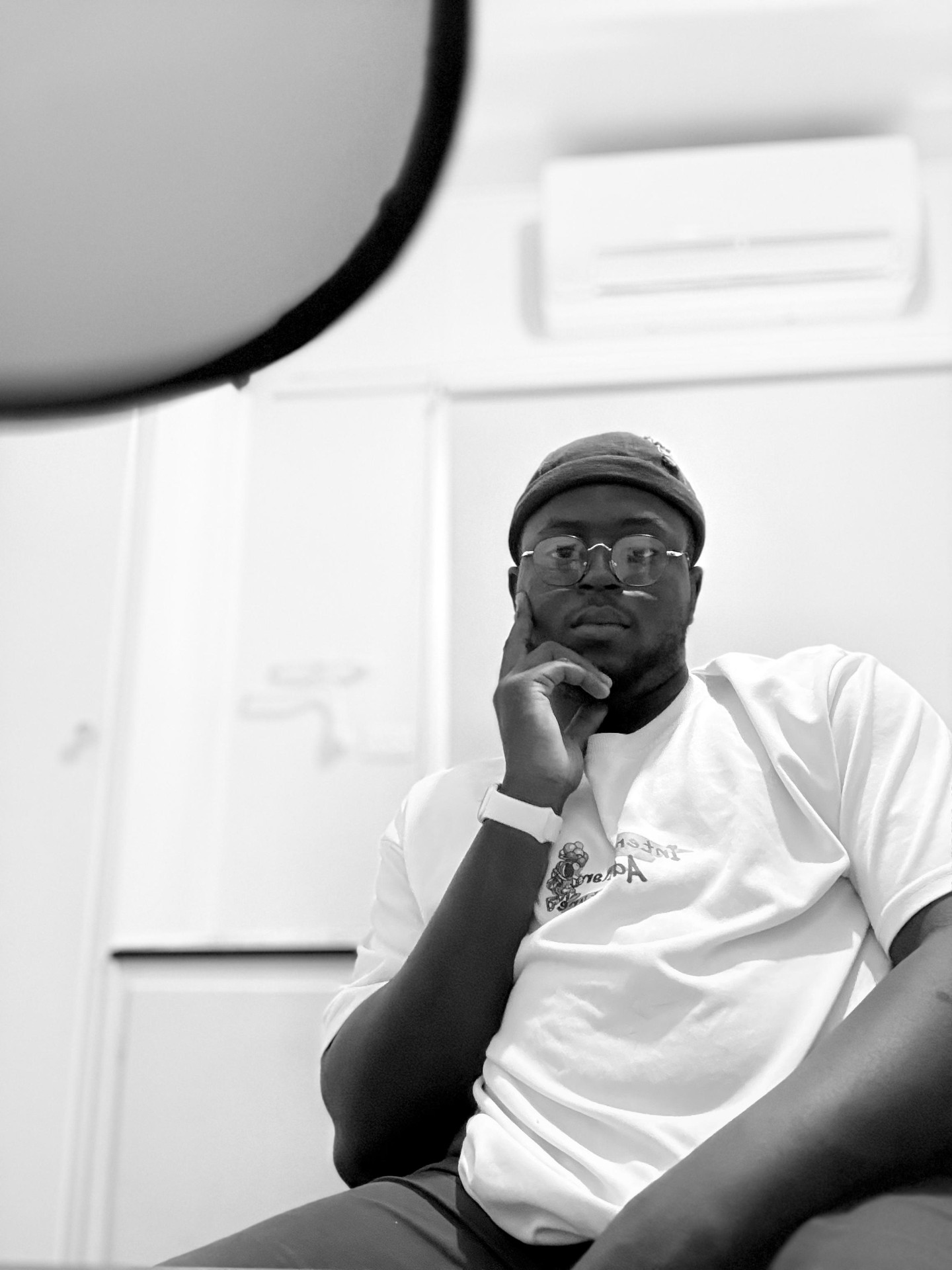 Black and white portrait of Oche, photographer, seated with a beanie and round glasses