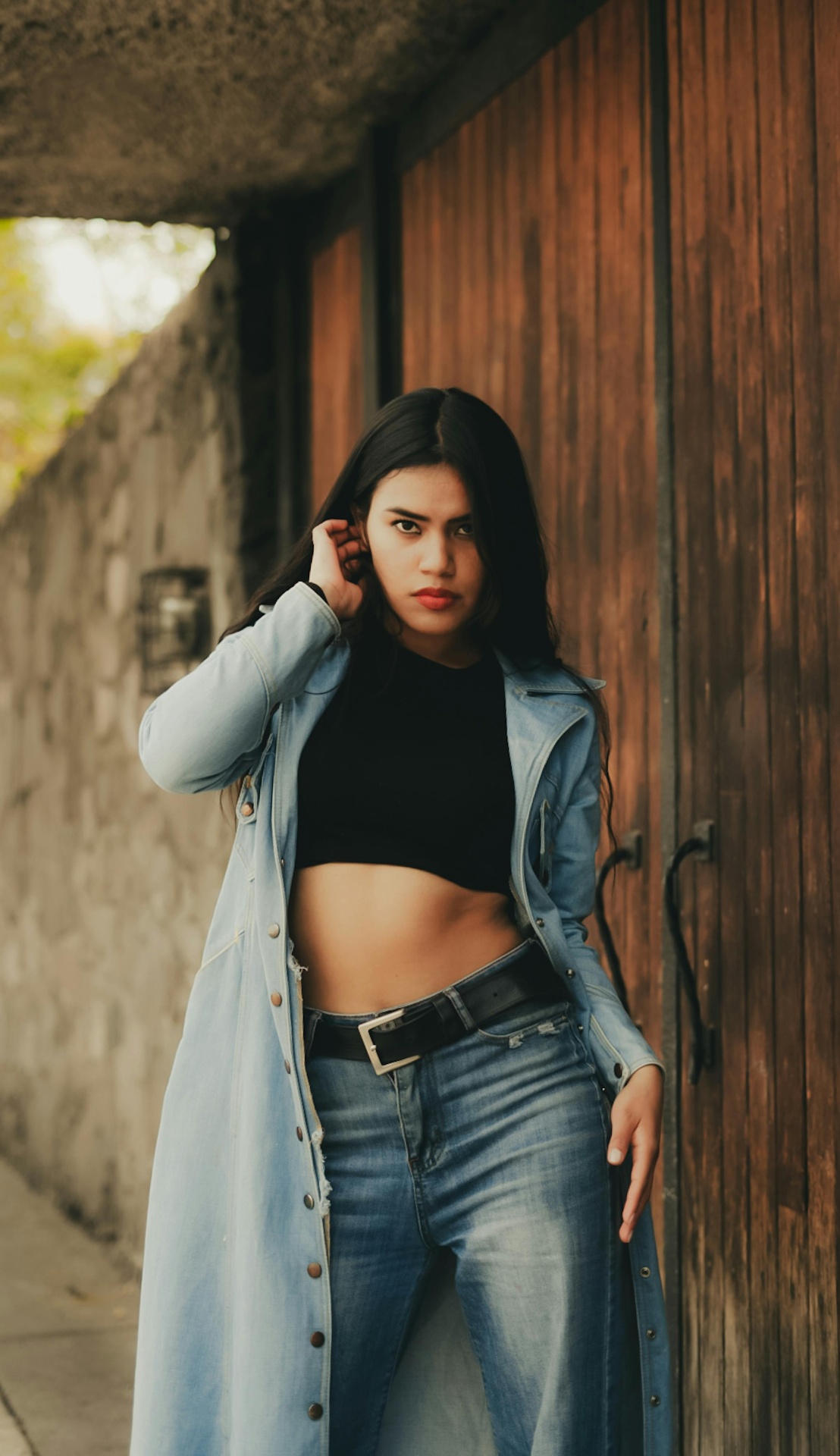 Street style portrait of a woman in a long denim coat leaning against a wooden door