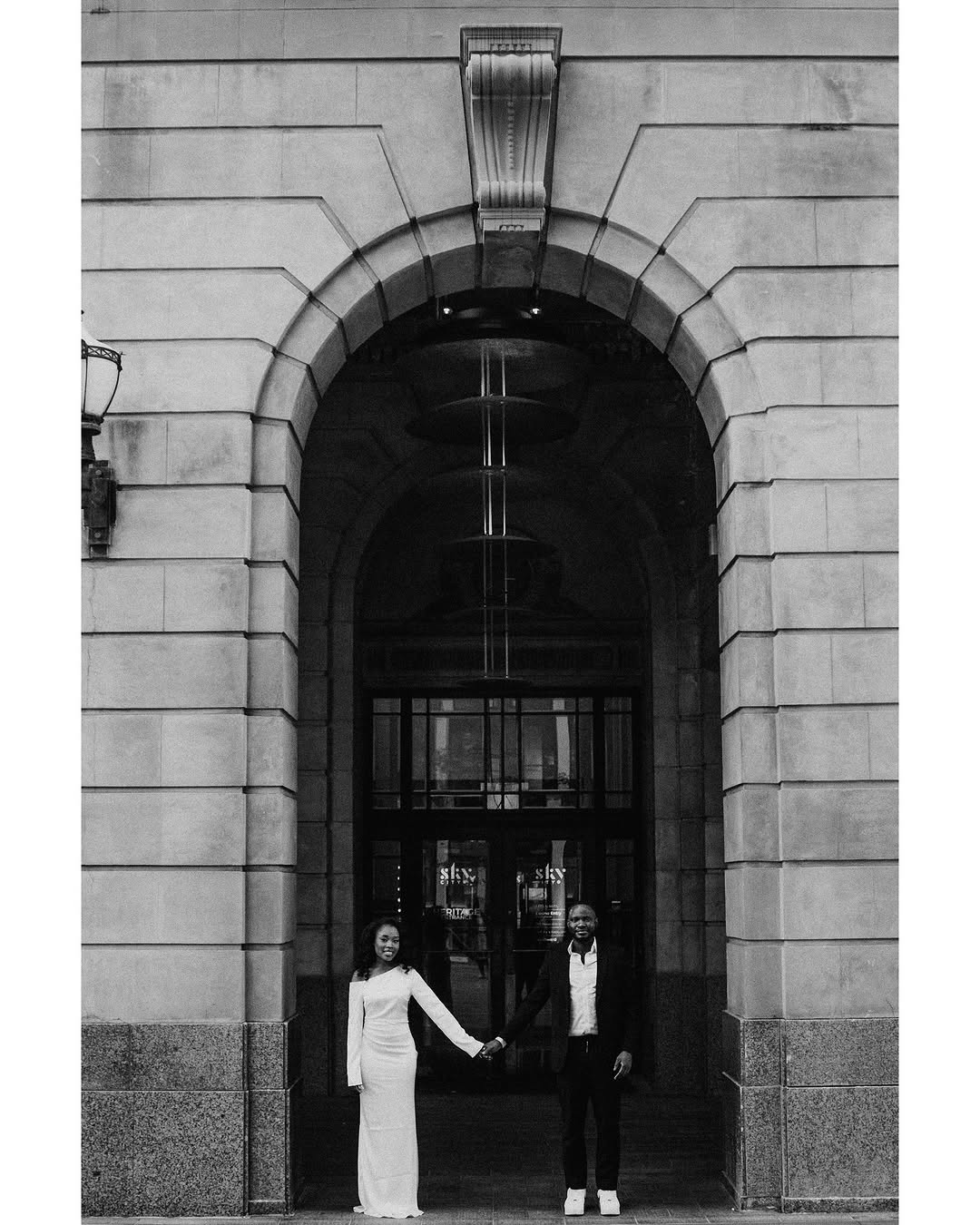 Black and white engagement portrait of couple holding hands beneath a grand stone archway